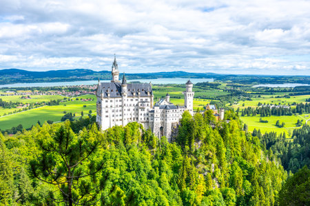 Neuschwanstein Castle rises beautifully from a green hillside in Bavaria, Germany, surrounded by lush forests and distant mountains under a vibrant sky, creating a fairytale ambiance.の写真素材