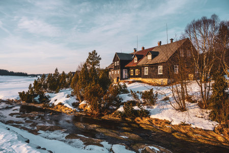 Bright sunlight bathes Jizerka village in winter. Snow covers the ground and rooftops, while a gentle stream flows by cottages nestled among trees, creating a serene, picturesque scene.の写真素材