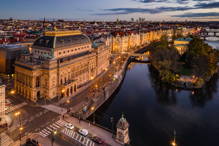 Aerial view of the National Theatre in Prague during evening time. The building is illuminated, with the Vltava River and embankment visible. The city lights reflect off the water.の写真素材