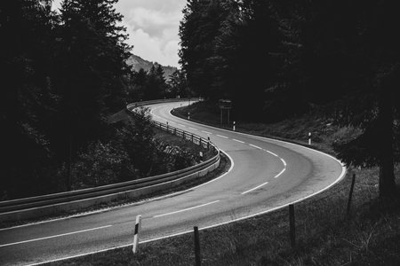 A winding road stretches through a lush green landscape, surrounded by dense trees and a cloudy sky. The road appears empty, inviting exploration and travel.の写真素材