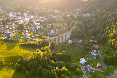 A railway bridge spans a green valley in Smrzovka. The structure connects areas, surrounded by trees and houses, during bright daylight hours.の写真素材