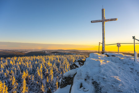 A cross stands on the summit rock of Jizera Mountain. Snow covers the ground and trees. Sunset casts warm light over the landscape, creating a striking contrast with the winter scene.の写真素材