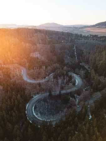 A winding road snakes through a lush forest as the sun sets, casting a warm glow over the landscape....