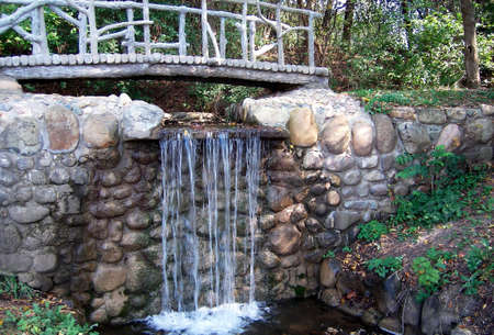 A small, artificial waterfall with a footbridge crossing over it in a park setting.の写真素材