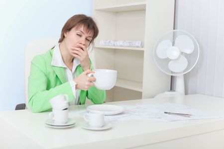 The tired woman sits at a table with the big cup of coffee and yawnsの写真素材