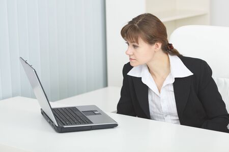 The young serious woman sits at a table at light office with the laptopの写真素材