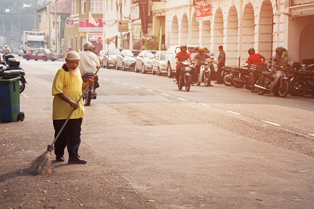 MALAYSIA, PENANG, GEORGETOWN - CIRCA JUL 2014: Old man sweeping the streets with a coarse, handmade broom as light traffic passes on the roadway.のeditorial素材
