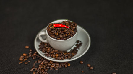 aromatic coffee beans in white cup with decoration of acute red chili peppers, spilled coffee beans,...