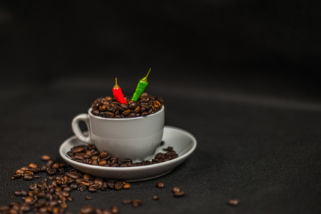aromatic coffee beans in white cup with decoration of acute red and green chili peppers, spilled coffee beans, artistic decorationの写真素材