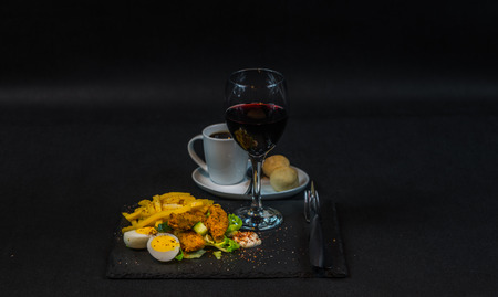 dish consisting of chicken breast breaded, French fries on ice lettuce with two halves of eggs, red wine in a glass and coffee, a dish on a black stone plate, lunch or dinnerの写真素材
