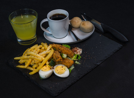 left side view on a dish consisting of chicken breast breaded, French fries on ice lettuce with two halves of eggs, black coffee and lemon juice, a dish on a black stone plate, lunch or dinnerの写真素材