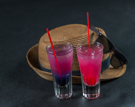Two colored drinks, a combination of dark blue with purple, the second combination of red with pink, long drink glass, brown sunglasses, a brown hat, party setの写真素材