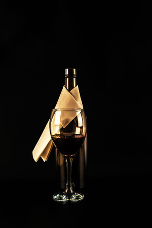 Open bottle of wine with white napkin, large glass filled with red wine, black background, elegant setの写真素材
