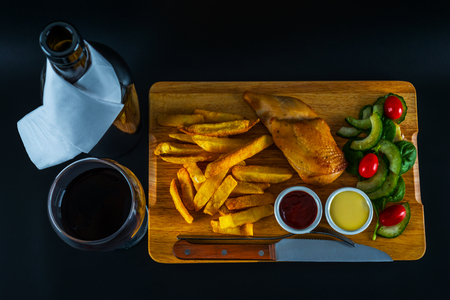 Roast chicken breast with chips, salad with tomatoes and cucumbers, wooden board, knife and fork, black backgroundの写真素材