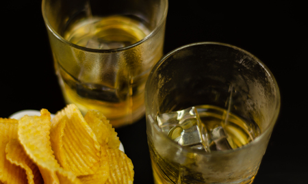 Glass of whiskey with ice cubes and salty snacks on a black background, party timeの写真素材