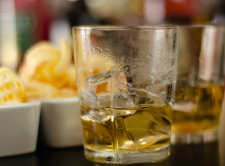 Glasses of whiskey with ice cubes and salty snacks on the background of bottles and bar, party timeの写真素材
