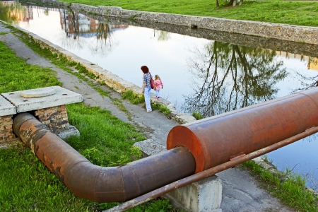 Woman and little girl going by waters bank. Large rusty metal pipe over the reservoir. View from the topの写真素材