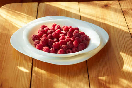 Ripe red raspberries in a plate on the wooden table partially lighted by sunlightの写真素材