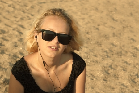 Beautiful young blond girl with glasses and headphones on the background of a sandy beach, edited with sepiaの写真素材