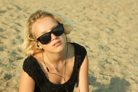 Beautiful teenage blond girl with glasses and headphones on the background of a sandy beachの写真素材