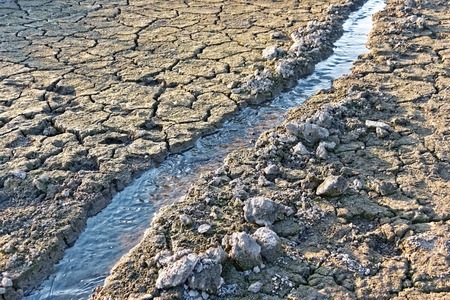 Water stream flowing in the channel among dried grey cracked soil in hot summerの写真素材
