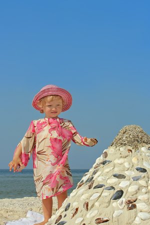 Beautiful little two-year girl standing on a sea beach near the sandy pyramid with shells on the background of a blue cloudless skyの写真素材