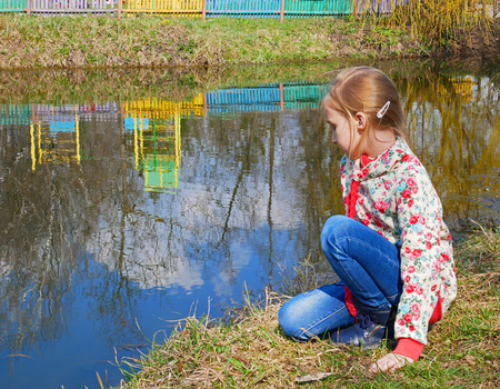 Small beautiful Caucasian girl sits near the lake in the park in springtimeの写真素材