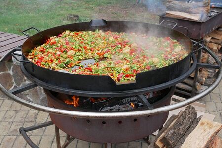 A lot of various multicolor sliced vegetables are fried in big handmade metal pan in boiling oil outdoorsの写真素材
