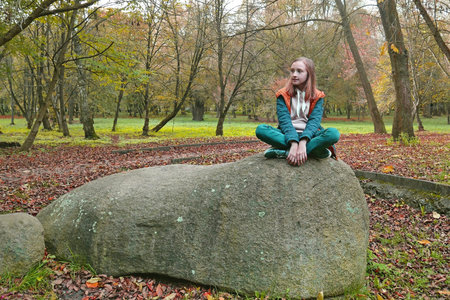 School age girl in turquoise and orange clothes sits on a huge shapeless boulder in an old autumn park, color editing photoの写真素材