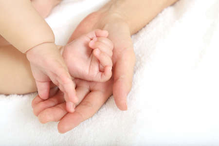 Mother's hand gently wrapping her baby's hand on a white sheetの写真素材