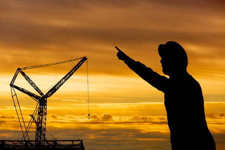Silhouette of crane and worker towering over construction site against sunset backgroundの写真素材