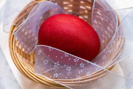 A red painted Easter egg lies in a wicker basket with a light blue ribbon on a white backgroundの写真素材