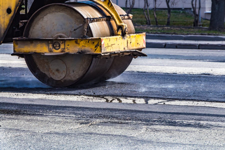 A road repair and an old rusty yellow roller rolls out a thin layer of a new black fuming hot...