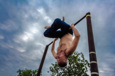 A man with a bare torso in blue pants upside down doing his morning exercises on a horizontal bar against the blue sky with clouds in the courtyard in the summer.の写真素材