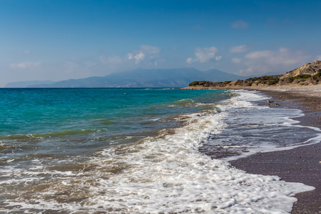 A beautiful deserted sand and pebble beach with foamy waves surf and mountains and a blue sky with white clouds are on the photoの写真素材