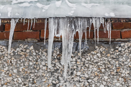 A group of sharp white transparent icicles is hanging down from the gray roof with ice of a building...