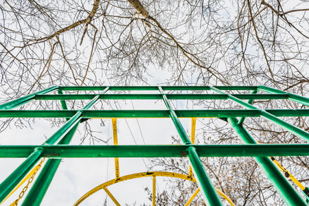 Green and yellow metal climbing grid against blue sky with white clouds and poplar and maple tree...