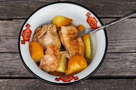 Juicy roast meat with golden potatoes served in a white metal bowl with a red ornament with a fork on an old rough gray wooden background.の写真素材