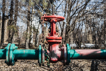 Red metal gas valve with a chain is on a green pipeline on a spring park background