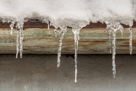 A group of sharp white and red transparent icicles is hanging down from the roof of a building in winterの写真素材