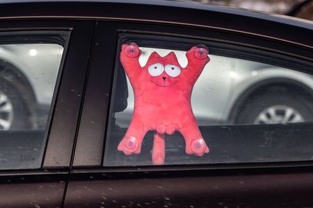 A beautiful little plush soft red toy cat with white eyes and a smile is hanging on the window inside a dirty carの写真素材