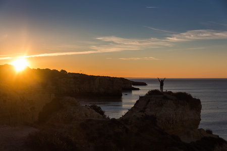 Rear view of a woman standing on the rock with raised arms and meeting the sun on the Atlantic Ocean in Portugalの写真素材