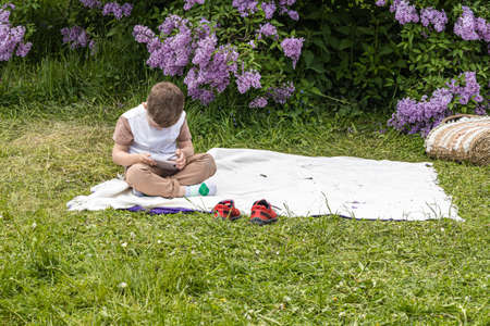 Yekaterinburg, Sverdlovsk / Russia - 06 06 2020: One Boy with a smartphone sits in the park on a white carpet against a background of purple lilacのeditorial素材