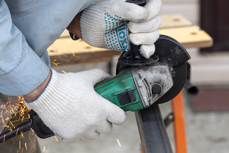 A man in the safety gloves is cutting metal using a green angle grinder tool with orange sparks on a...