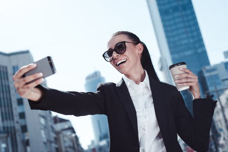 It is selfie time! Beautiful young businesswoman in sunglasses making selfie with smile outdoors.の写真素材