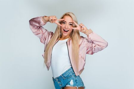 Peace to you! Beautiful young woman with blond hair looking at camera and gesturing peace with smile while standing against gray backgroundの写真素材