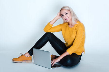 Beautiful young woman with blond hair using her laptop and looking at camera with smile while...