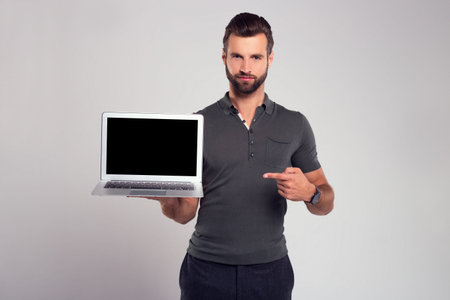 Your new device. Handsome young man looking at camera and pointing at his laptop while standing...