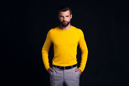 Simple and stylish. Handsome young man looking at camera and keeping hands in pockets while standing against black backgroundの写真素材