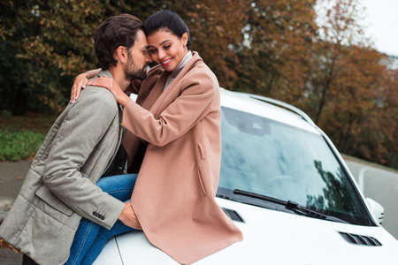 Sensual autumn trip is about to begin. Young beautiful couple bonding with smile while standing outdoors with the car at the backgroundの写真素材
